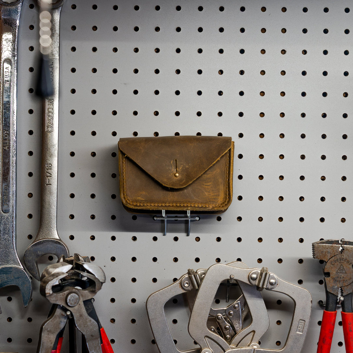 Brown leather Standard Issue Leather Case hanging on a pegboard wall above wrenches and pliers